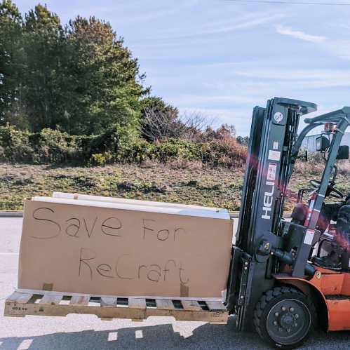A forklift carrying a large cardboard box with "Save For ReCraft" written on it, set against a natural background.