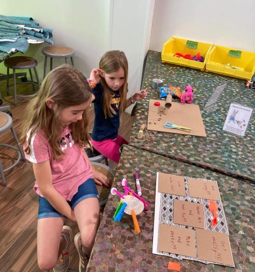 Two young girls are engaged in a craft activity at a table, surrounded by various art supplies and materials.