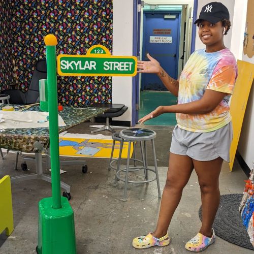 A person in a tie-dye shirt and shorts stands next to a green street sign that reads "SKYLAR STREET."