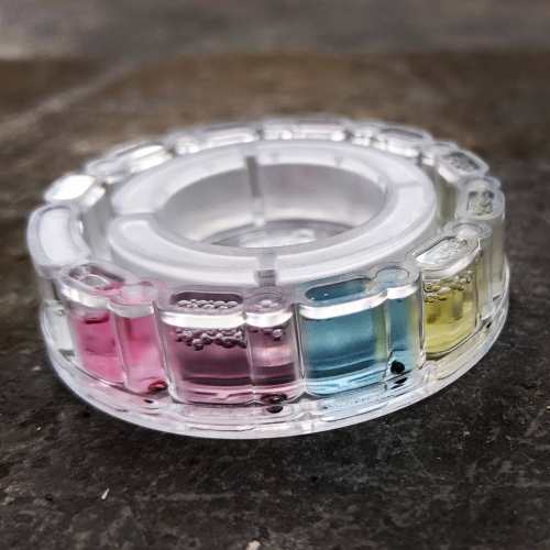 A circular clear container with colored liquids in segments: pink, blue, yellow, and clear, resting on a dark surface.