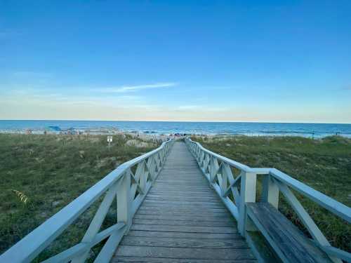 A wooden boardwalk leads to a sandy beach under a clear blue sky, with gentle waves in the distance.