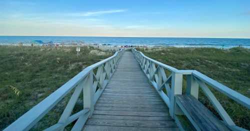 A wooden boardwalk leads to a sandy beach with gentle waves and people in the distance under a clear blue sky.