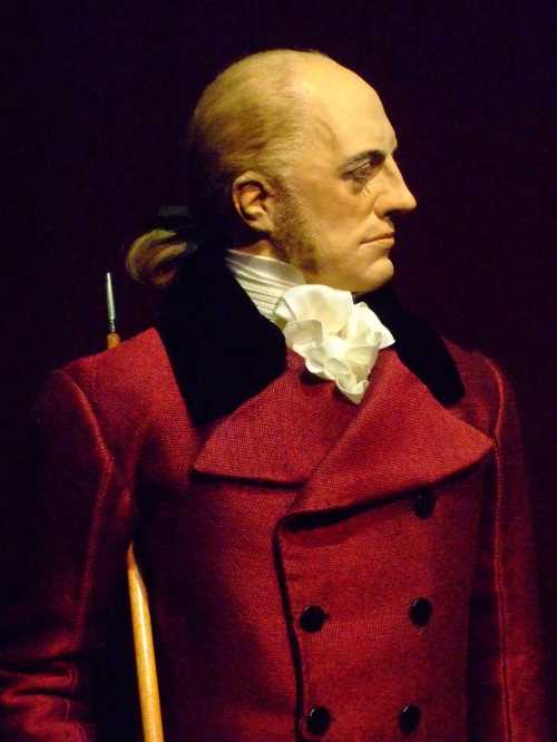 A historical figure in a red coat with a white cravat, looking to the side, with a serious expression.