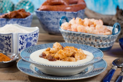 A bowl of shrimp and grits, surrounded by bowls of shrimp, sausage, and spices on a rustic table.