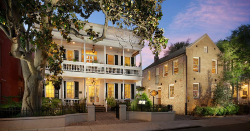 Historic buildings with balconies and large windows, surrounded by trees, illuminated at dusk.