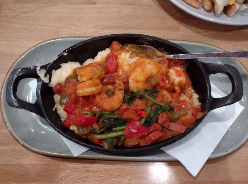 A cast iron dish with shrimp, tomatoes, spinach, and spices served over creamy grits on a wooden table.