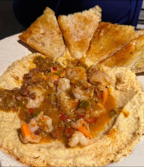 A plate of hummus topped with shrimp and spices, served with toasted bread slices arranged around it.