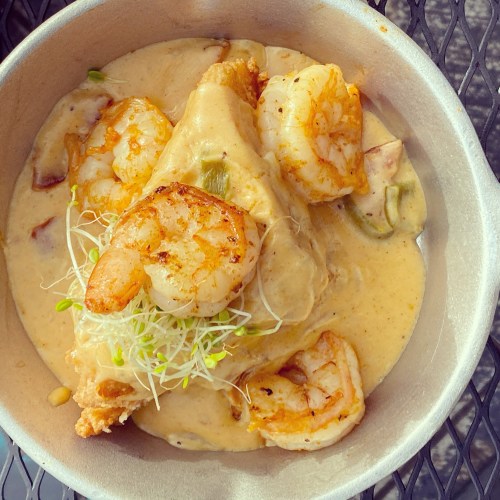 A bowl of creamy shrimp sauce over a fried fish fillet, garnished with sprouts and served in a metal dish.