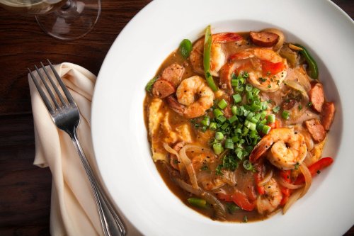 A bowl of shrimp and sausage gumbo topped with green onions, served with a fork and napkin on a wooden table.