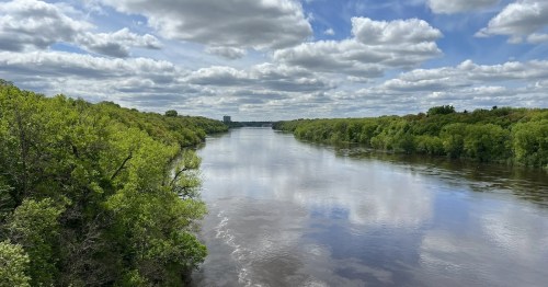 A serene river flows through lush green trees under a cloudy blue sky.