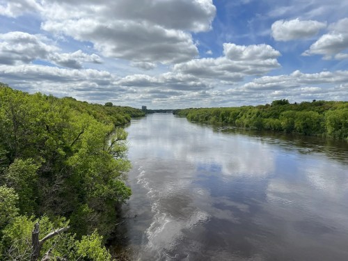 A serene river flows through lush greenery under a partly cloudy sky, reflecting the clouds on its surface.