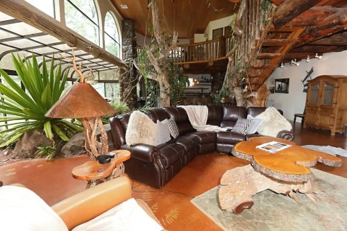 Cozy living room with a brown sectional sofa, wooden furniture, and large windows surrounded by greenery and natural elements.