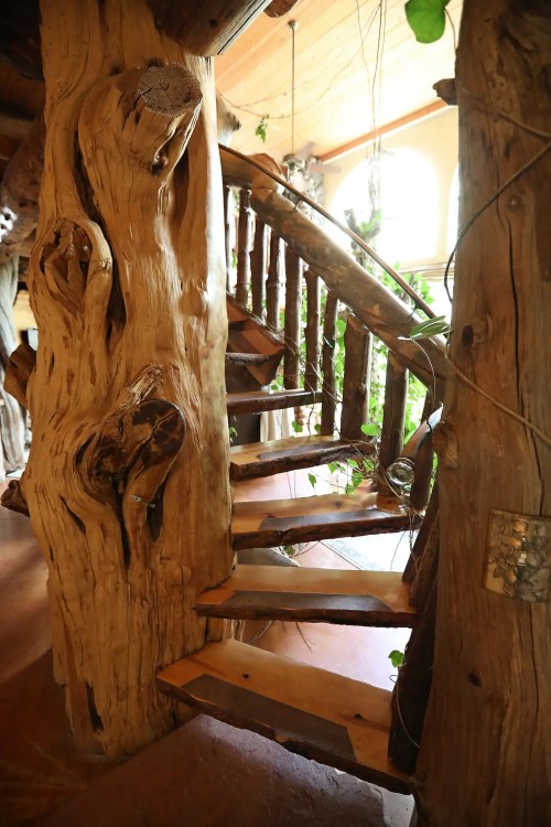 A rustic wooden staircase winds up through a natural wood structure, surrounded by greenery and warm lighting.