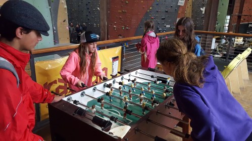 A group of children play foosball indoors, with a climbing wall visible in the background.