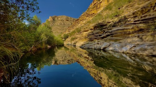 A serene river flows between rocky cliffs, reflecting the blue sky and surrounding greenery.