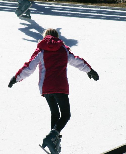 A child in a red jacket ice skating on a frozen surface, with other skaters in the background.