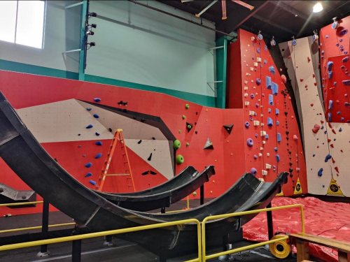 Indoor climbing gym featuring colorful climbing walls, a ladder, and curved climbing surfaces.