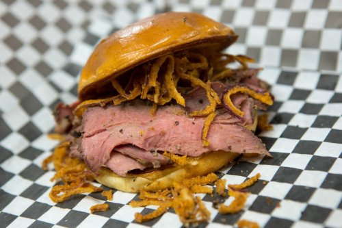 A close-up of a sandwich with sliced roast beef and crispy onions on a soft bun, served on checkered paper.