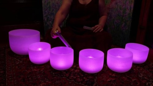 A person plays glowing purple crystal singing bowls on a patterned rug, creating a serene atmosphere.