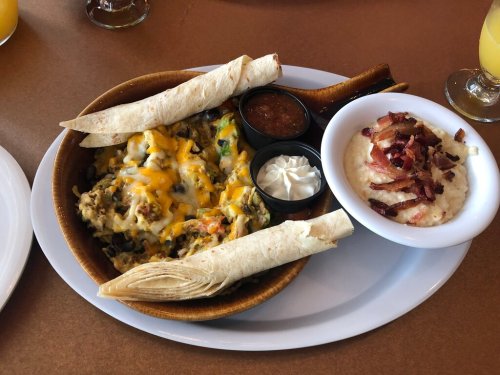 A bowl of cheesy nachos with toppings, two rolled tortillas, and a side of creamy grits topped with bacon bits.