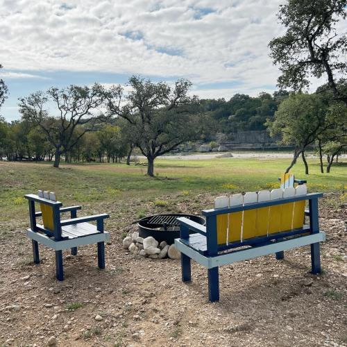 Two colorful benches face a fire pit in a serene outdoor setting with trees and a grassy area in the background.