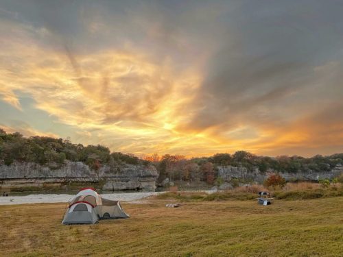 A tent sits on grassy land with a stunning sunset and colorful clouds in the background, surrounded by trees.