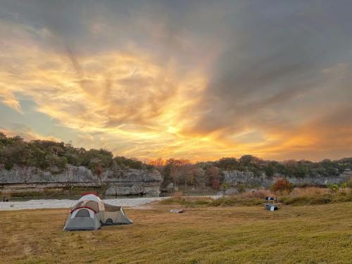 A tent sits on grassy ground with a stunning sunset and colorful clouds in the background, surrounded by trees.