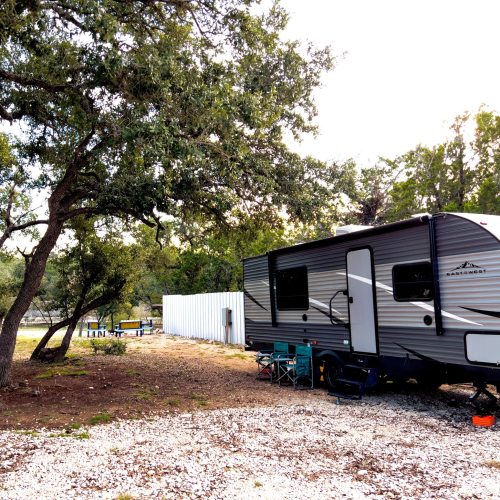 A parked RV beside a tree, with camping chairs set up nearby in a natural outdoor setting.