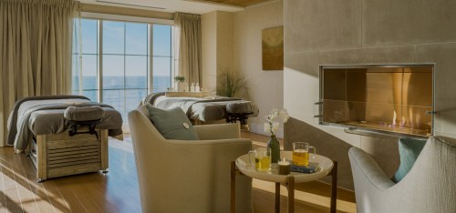 A serene spa room with two massage tables, a cozy seating area, and a view of the ocean through large windows.