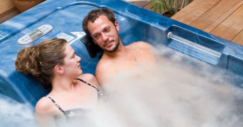 A couple relaxes in a hot tub, surrounded by steam and a wooden deck, enjoying each other's company.