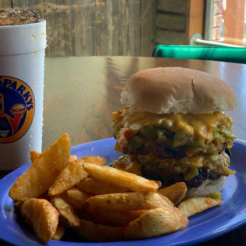 A stacked burger with cheese and toppings, served with potato wedges and a drink in a cup on a wooden table.