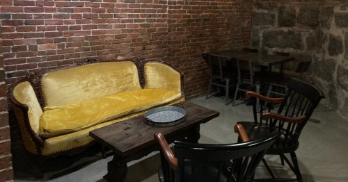 Cozy, rustic room with a yellow velvet couch, wooden table, and black chairs against a brick and stone wall.