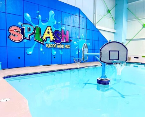 Brightly colored "SPLASH" mural on a wall beside a clear pool with basketball hoops in an indoor water park.