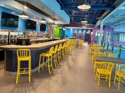 Brightly lit bar area with yellow chairs, modern decor, and multiple TVs in a spacious venue.