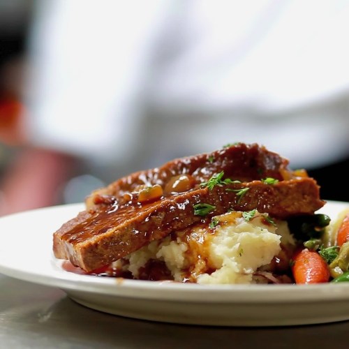 A plate of meatloaf topped with sauce, served with mashed potatoes and green garnish, on a restaurant table.