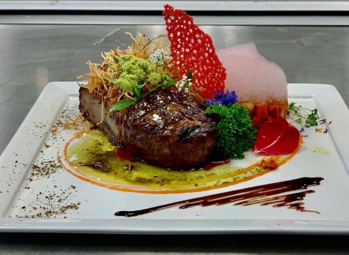 A beautifully plated steak with garnishes, colorful foam, and decorative elements on a white dish.