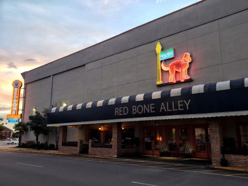Exterior of Red Bone Alley restaurant with a neon dog sign and awning, set against a colorful sunset sky.