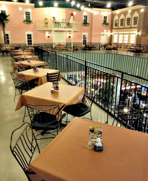 A spacious restaurant interior with empty tables, warm lighting, and a balcony overlooking a lively dining area.