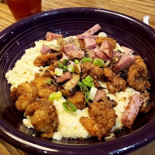 A bowl of creamy grits topped with fried chicken, diced ham, mushrooms, and green onions.