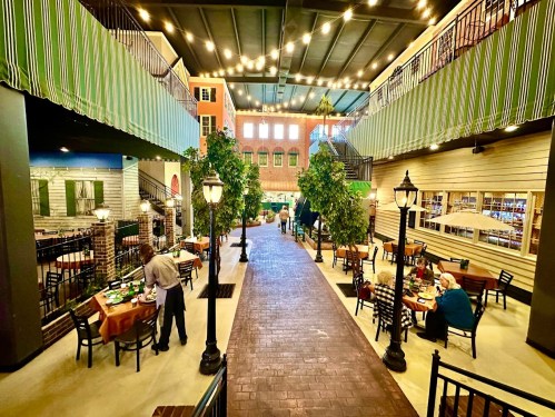 A vibrant indoor dining area with tables, greenery, and decorative lighting, resembling a charming street scene.