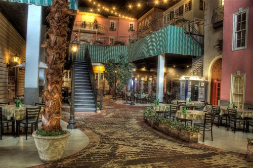 A vibrant indoor courtyard with brick pathways, palm trees, and dining tables under string lights and colorful buildings.