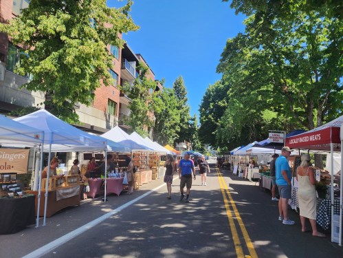 A vibrant street market with white tents, vendors, and shoppers under clear blue skies and green trees.