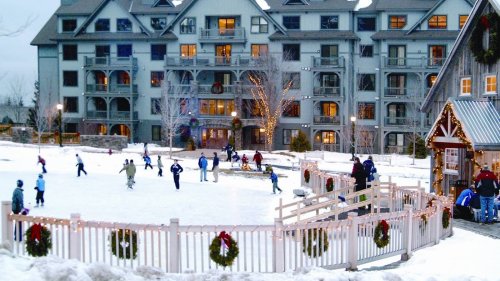 A winter scene with people ice skating on a rink outside a lodge, decorated with holiday lights and wreaths.