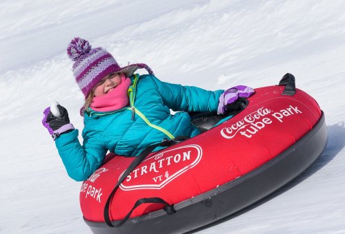 A child in a colorful winter jacket enjoys tubing on snow, smiling and giving a thumbs up while sitting in a red tube.