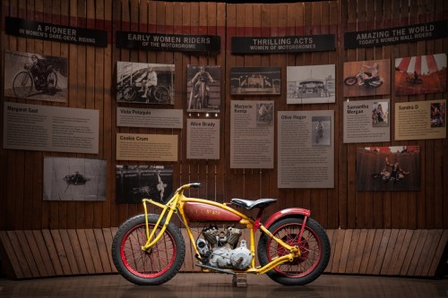 A vintage motorcycle displayed in front of a wall featuring historical photos and information about women motorcyclists.