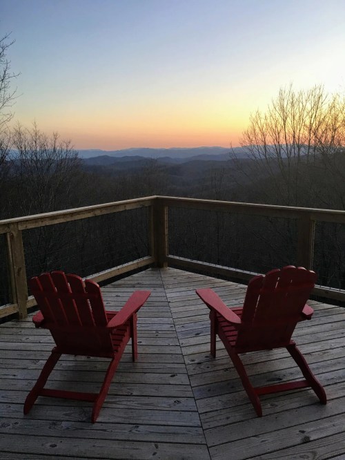 Two red Adirondack chairs on a wooden deck overlook a serene mountain landscape at sunset.
