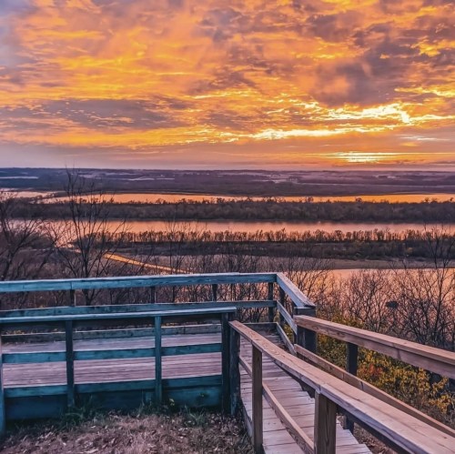 A wooden overlook with a view of a river and vibrant sunset sky filled with clouds.