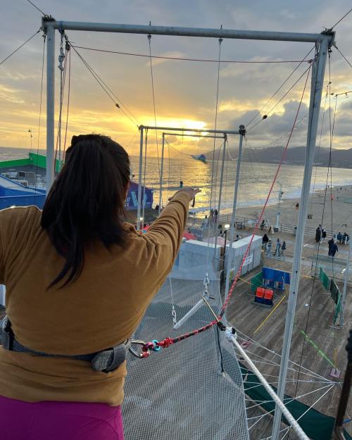 A person in a harness points towards a sunset over the ocean, with a trapeze setup in the foreground.