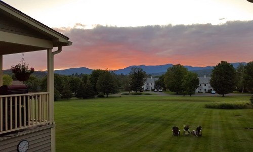 A serene sunset over mountains, with a green lawn and chairs in the foreground, viewed from a balcony.