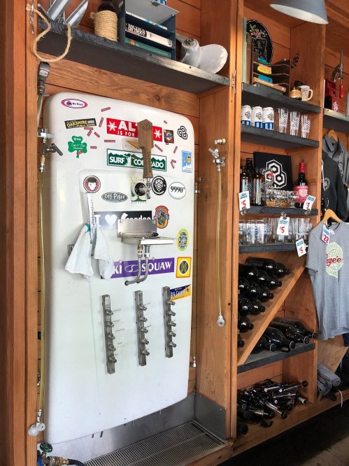 A vintage fridge covered in stickers, with taps for drinks, surrounded by shelves of bottles and merchandise.